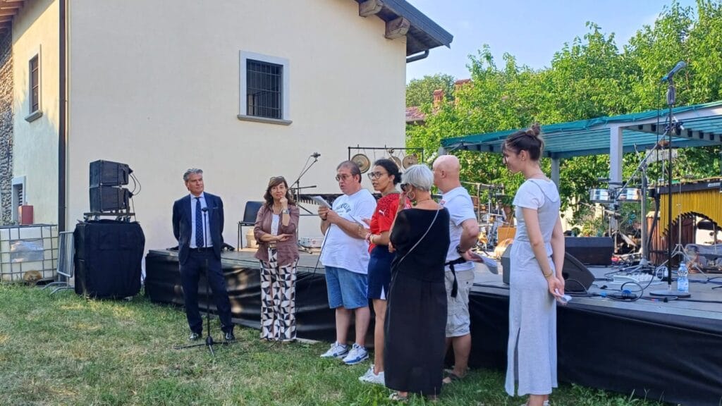 Presentazione del progetto "Storie di quartiere" durante la Festa dei Vicini 2026 a Boccaleone: un gruppo di partecipanti prende la parola davanti al palco, con il pubblico riunito nel cortile della biblioteca per condividere l'esperienza di raccolta delle storie di vita dei residenti del quartiere.