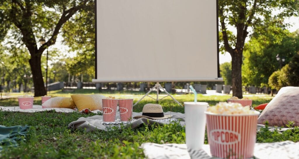 Schermo per cinema all’aperto allestito in un parco, con teli e cuscini sull’erba e contenitori di popcorn.