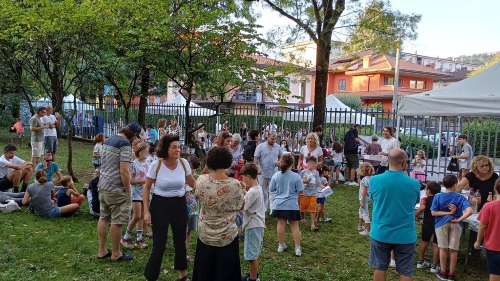 Gruppo di adulti e bambini riuniti in un’area verde del quartiere durante una festa all’aperto, tra momenti di incontro, attività e convivialità.